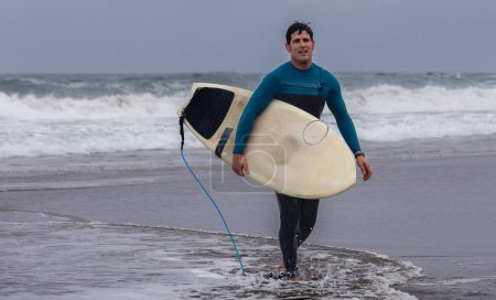 Foto de Un hombre surfista cansado con un traje azul sale del océano llevando su tabla de surf después de una sesión. - Imagen libre de derechos