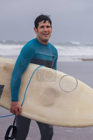 Téléchargez les photos : Un homme souriant en combinaison bleue tient une planche de surf, prête pour les vagues par une journée nuageuse à la plage. - en image libre de droit