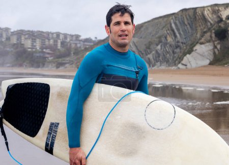 Un surfeur masculin en combinaison bleue se tient à côté de sa planche de surf avec des maisons côtières et des falaises en arrière-plan.
