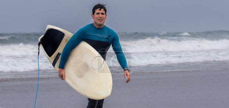 Foto de Amplio tiro de un surfista en un traje de neopreno azul llevando su tabla de surf a lo largo de la orilla, con olas estrellándose en el fondo. - Imagen libre de derechos