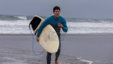 Ein aktiver Surfer im blauen Neoprenanzug rennt mit seinem Surfbrett am Strand entlang, um die Wellen zu fangen.