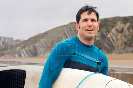Fröhlicher Mann im Krickenten-Neoprenanzug hält ein Surfbrett an einem Strand mit markanten Klippen hinter sich.