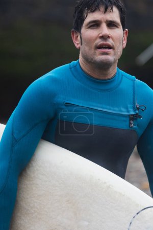 Gros plan d'un surfeur en combinaison bleue tenant sa planche de surf, avec un regard concentré après le surf.