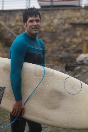Ein Surfer lächelt optimistisch und hält sein Surfbrett an einer Meereswand, nachdem er die Wellen des Ozeans genossen hat.