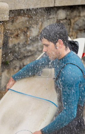 A surfer, holding his board, enjoys the refreshing droplets of an outdoor shower, washing off the ocean's embrace