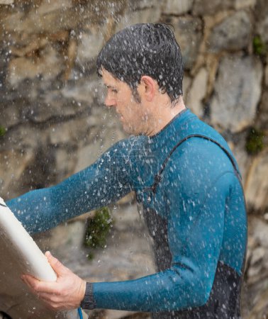 Téléchargez les photos : Un surfeur concentré en combinaison bleue nettoie méticuleusement sa planche de surf sous une douche à la plage. - en image libre de droit