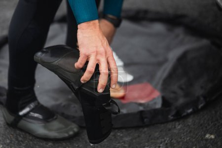 Close-up of a surfer's hands adjusting neoprene wetsuit boots on a mat by the beach.