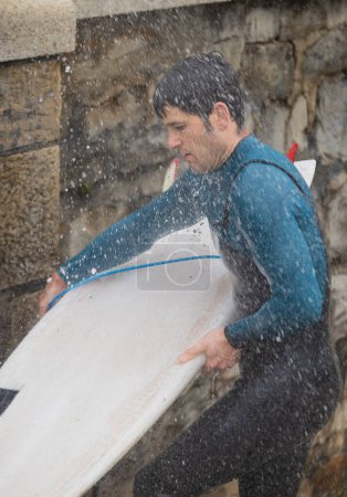 Engrossi dans sa routine, un surfeur lave sa planche sous une douche extérieure, l'eau éclaboussant autour de lui.