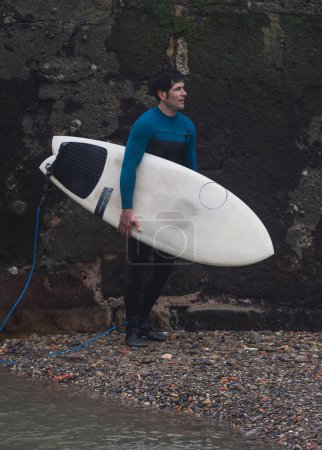 A male surfer stands by a rugged shore, holding a surfboard, contemplating the waves.