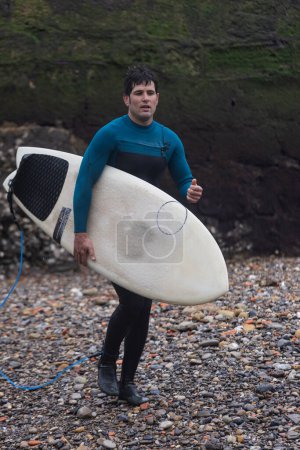 Téléchargez les photos : Un surfeur masculin se tient près d'un rivage rocheux, tenant une planche de surf, prêt pour une séance de surf. - en image libre de droit