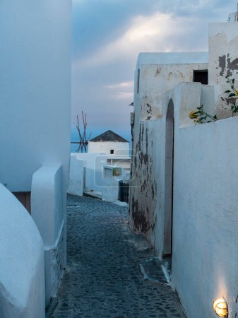 A quaint Santorini alleyway leads to a windmill, captured in the soft glow of dusk, embodying the essence of Greek island charm.