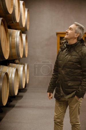 A thoughtful winemaker stands among oak barrels, appreciating the aging process of fine wine.