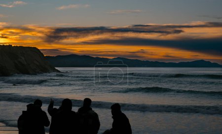 Silhouetten von Freunden zusammen am Strand und beobachten die Wellen unter einem pulsierenden Sonnenuntergang.