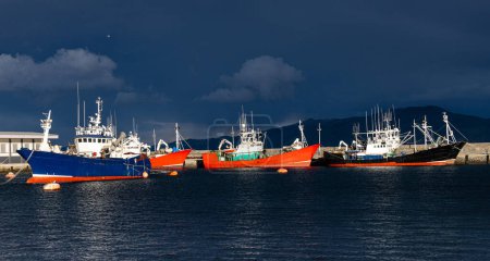 Foto de Coloridos barcos de pesca anclados en el puerto con un cielo tormentoso dramático en el fondo, capturando una serena escena marítima. - Imagen libre de derechos