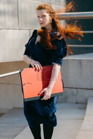A poised red-haired woman in a navy dress holds a red portfolio, her hair swept by the wind against an architectural backdrop.