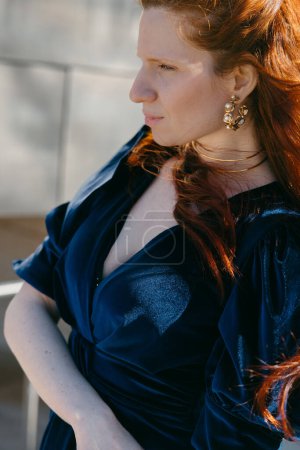 Eine kontemplative Frau mit feuerroten Haaren trägt ein luxuriöses Samtkleid vor einem eleganten metallischen Hintergrund..