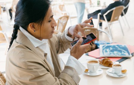 Eine fokussierte junge Berufstätige benutzt ihr Smartphone in einem Café, umgeben von einer frischen Tasse Kaffee und Gebäck.
