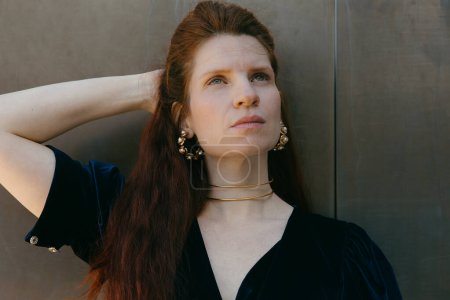 A woman with long red hair, adorned with stylish jewelry, looks off into the distance, deep in thought.