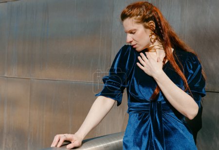 Un estado de ánimo reflexivo capturado como una mujer en un vestido de terciopelo azul toca su pecho, de pie junto a una pared de metal.