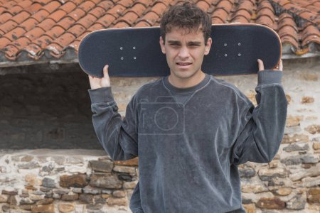 Portrait of a determined young male skater holding his skateboard behind his head against a rustic background, showcasing urban lifestyle.