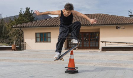 Photo for A skateboarder in a tank top jumps over a traffic cone, showcasing precision and control in an urban setting. - Royalty Free Image