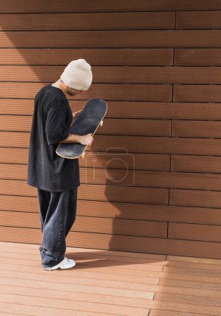 Photo for A reflective skateboarder holds his board close, standing by a wooden wall, in a moment of quiet contemplation. - Royalty Free Image