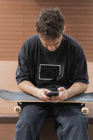 A skateboarder takes a break to check his smartphone, sitting on his skateboard on a wooden ramp.