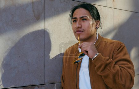 Young Ecuadorian man in a stylish brown jacket holding sunglasses, exploring the urban environment.