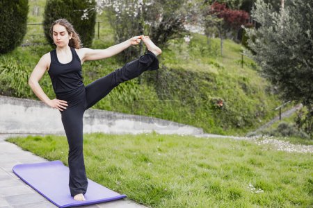 Téléchargez les photos : Une femme maintient une posture d'étirement de jambe de yoga difficile sur un tapis dans un cadre extérieur serein, mettant en valeur la flexibilité et l'équilibre. - en image libre de droit