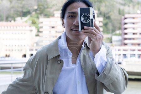 Téléchargez les photos : Outdoor portrait of a cheerful Ecuadorian man taking a photo with a retro-style camera in an urban environment - en image libre de droit
