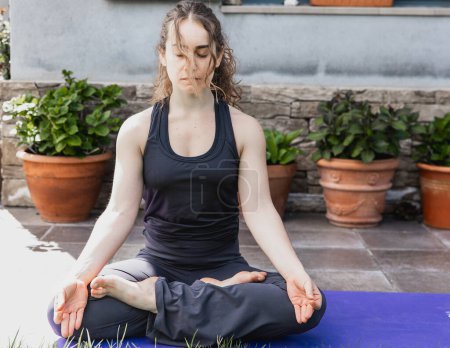A woman meditates in lotus position on a yoga mat, embodying tranquility in an outdoor setting.
