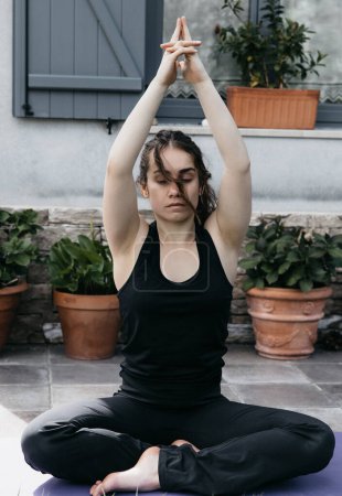A woman meditates in a seated prayer pose, eyes closed, with plants in the background.