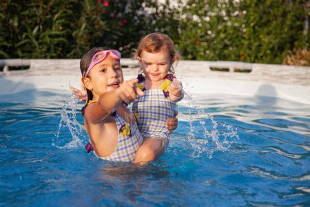 Téléchargez les photos : Deux jeunes sœurs éclaboussent d'eau en jouant dans une piscine, avec une végétation luxuriante en arrière-plan par temps clair. - en image libre de droit