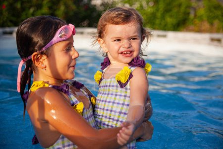 Two happy sisters smiling and having fun while playing together in a swimming pool, highlighted by a sunny garden backdrop.