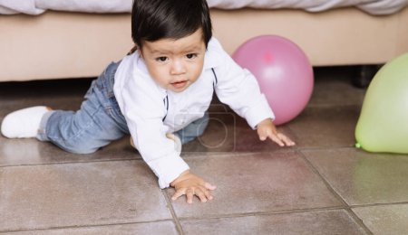 Foto de "Encantadora foto de un niño arrastrándose en un suelo de baldosas en casa, moviéndose hacia globos de colores. - Imagen libre de derechos