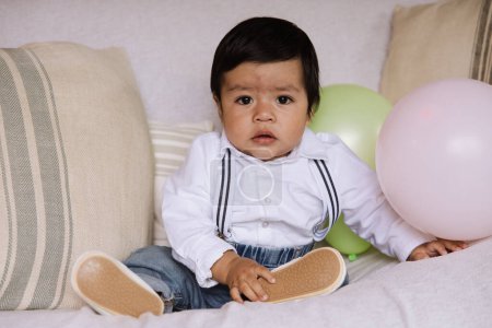 Portrait d'un mignon bébé garçon assis sur un canapé, vêtu d'une chemise blanche élégante avec des bretelles et un jean en denim.