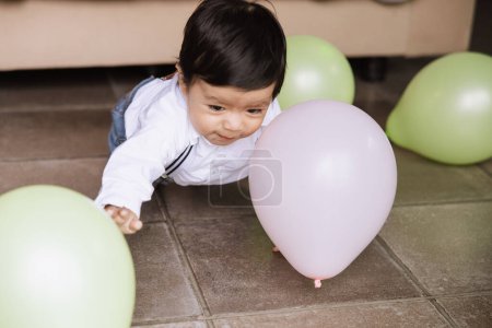 Foto de Foto cautivadora de un niño alegre arrastrándose sobre un suelo de baldosas, alcanzando un globo rosa entre otros. - Imagen libre de derechos