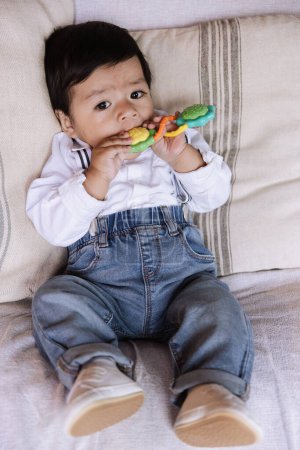 Foto de Adorable niño sentado en un sofá, vestido con una camisa blanca y vaqueros azules, masticando cómodamente un colorido juguete para la dentición. - Imagen libre de derechos