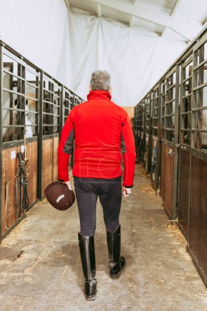 Vista trasera de un ecuestre caminando por el pasillo estable, sosteniendo un casco y una cosecha, listo para comenzar una sesión de equitación.