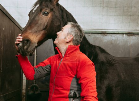 Photo for Cheerful man in a red jacket joyfully interacting with his dark brown horse in a stable, showcasing their strong bond and happiness. - Royalty Free Image