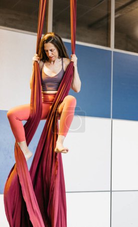 Una mujer practicando sedas aéreas en un estudio de gimnasia, trepando tela granate con concentración y fuerza. Ella está vestida con ropa deportiva, mostrando su habilidad y dedicación.