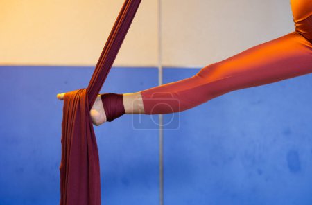 Close-up of a woman's leg wrapped in red fabric during aerial acrobatics training in a studio with blue and beige walls.