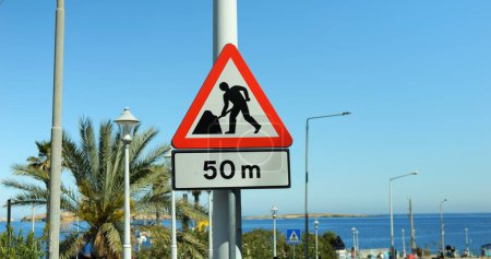 Photo for Road work sign with attached fifty meters distance plate mounted at lamp mast. Traffic sign warning about roadworks or construction ahead with sea and exotic palm tree in background in warm country. - Royalty Free Image