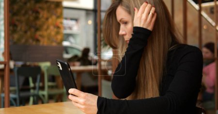 Stilvolle Frau in städtischen Coffeeshop verwenden Telefon-Bildschirm mit Spiegel-App und fixieren Haare. Modische weibliche Erwachsene sitzt allein am Tisch im Restaurant und achtet auf ihre Sicht. Hochwertiges Foto