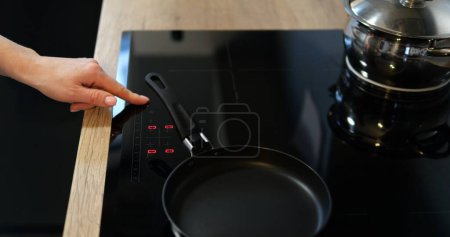 Foto de La mano femenina enciende la inducción negra o la encimera eléctrica con sartenes. Cómoda cocina equipada con un moderno y conveniente dispositivo de cocción. Imágenes de alta calidad 4k - Imagen libre de derechos