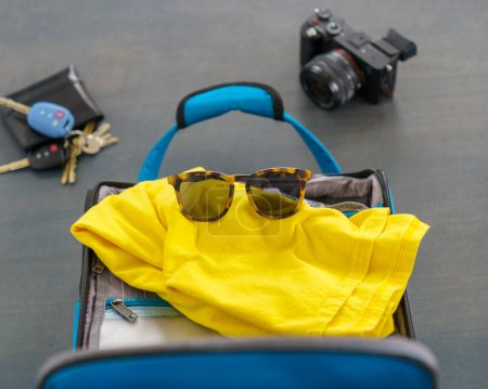Photo for Blue suitcase with a yellow shirt, sunglasses, camera, wallet, and keys. Ready for a vacation holiday getaway. - Royalty Free Image