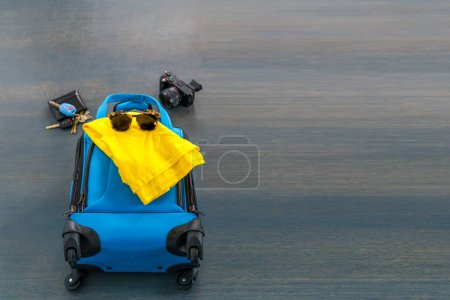Photo for Travel bag with a yellow shirt, sunglasses, wallet, keys, and a camera on a gray concrete floor. Ready for a vacation holiday. - Royalty Free Image