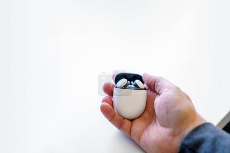Photo for Wireless earbuds headphones in their charging case with the lid open, being held by a caucasian adult male hand. White desk and room for copy space. - Royalty Free Image