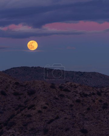 Vollmond steigt mit wolkenverhangenem Pastellhimmel über den Berggipfeln der Wüste im Yucca Valley, Kalifornien