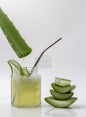 Aloe vera gel dripping on a jar of aloe vera juice with sliced and stacked aloe vera isolated on white background.